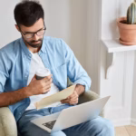 serious-entrepreneur-takes-care-business-works-home-concentrated-into-documents-has-laptop-computer-knees-holds-takeaway-coffee-sits-comfortable-armchair_273609-19041